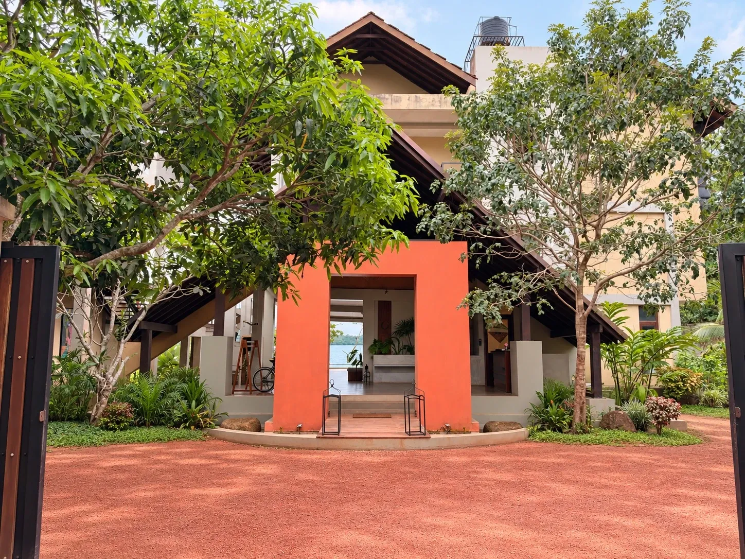 The entrance pavilion, framed in orange under a pitched roof, with trees either side of the red gravel drive (parked cars digitally removed)
