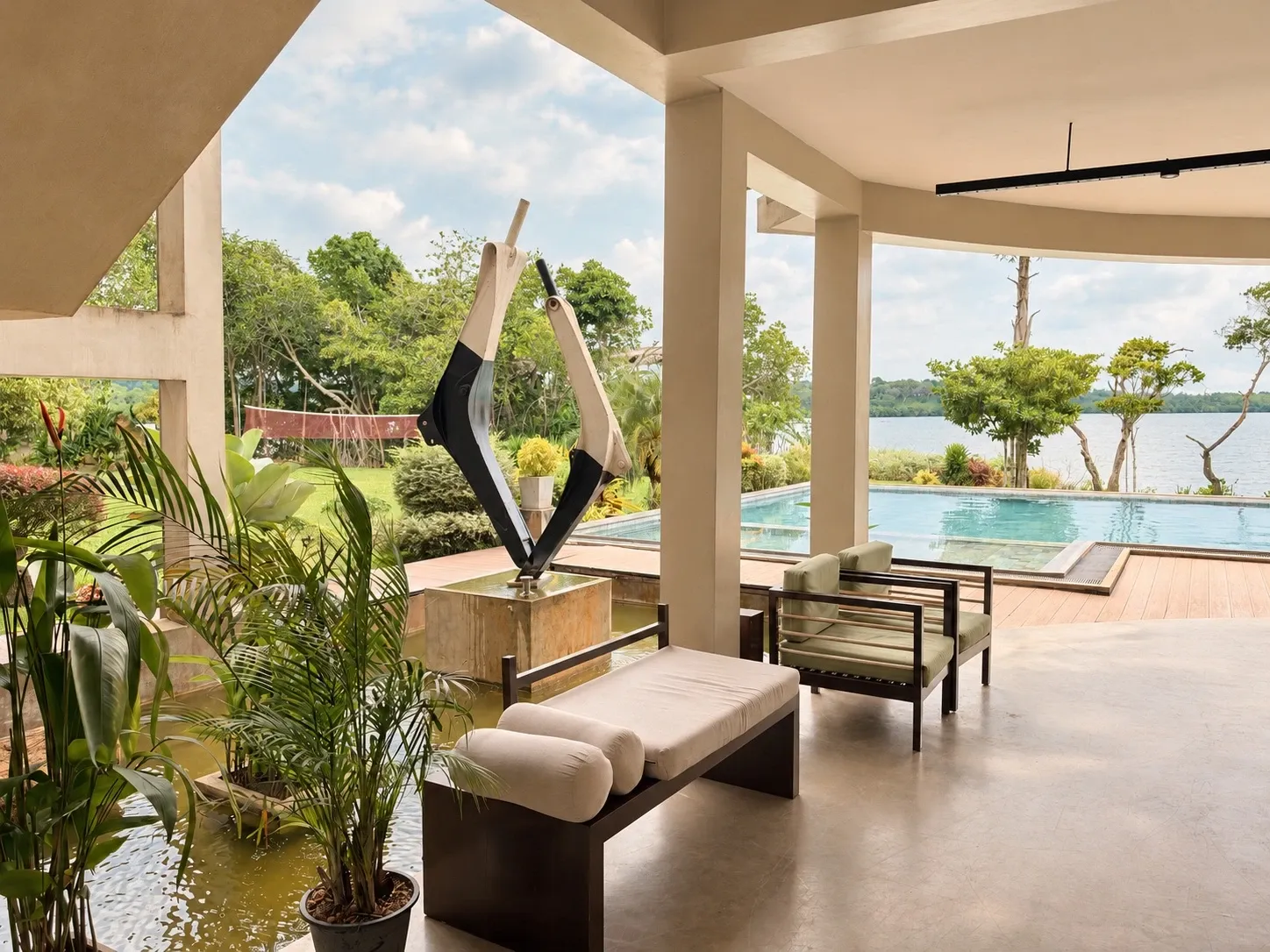 A covered terrace with a cushioned bench, potted palms and a black-and-white sculpture, looking over the pool to the lake